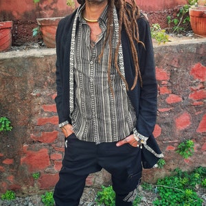 Op de afbeelding: Een persoon draagt een zwart vest met witte patroonafwerking, een overhemd met patroon en een zwarte broek. De persoon heeft dreadlocks en tatoeages. De achtergrond is een rode bakstenen muur met potplanten.