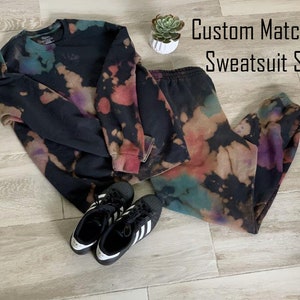 Può includere: Un set di felpa e pantaloni da tuta in tie-dye nero e bianco. Il set presenta un motivo tie-dye colorato con sfumature di viola, blu e rosso. Il testo "Custom Matching Sweatsuit Set" è visibile nell'immagine.