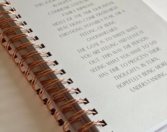 May include: Open journal with lined pages and a copper-colored spiral binding. The text on the page reads, "This journal is a simple tool in hopes to help communication between family improve."