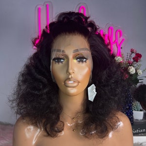 May include: A black curly human hair wig on a mannequin head. The wig has a lace front and is styled in a voluminous, curly look.