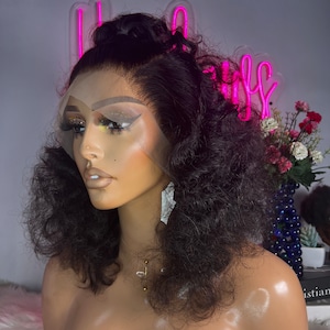 May include: A black curly human hair wig on a mannequin head. The wig has a lace front and is styled with a high ponytail. The mannequin is wearing a gold necklace.
