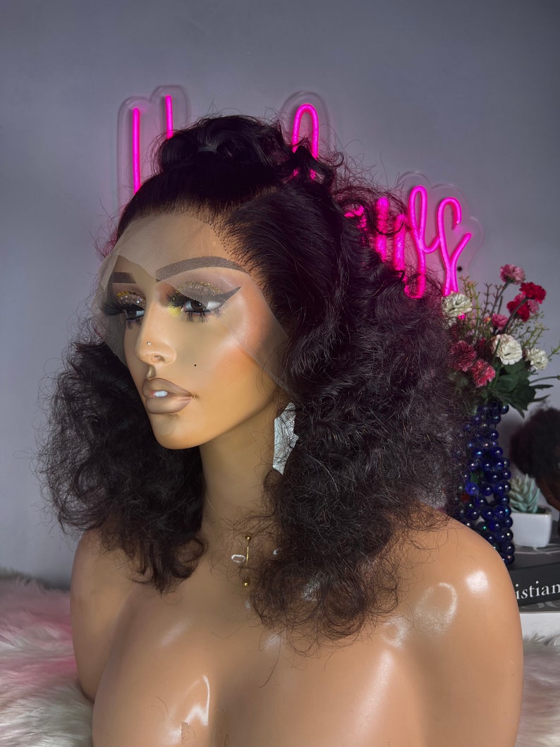 May include: A black curly human hair wig on a mannequin head. The wig has a lace front and is styled with a high ponytail. The mannequin is wearing a gold necklace.