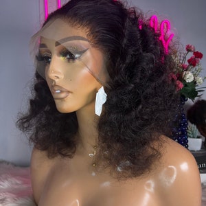 May include: A black curly human hair wig on a mannequin head. The wig has a lace front and is styled in a short, voluminous curly style.