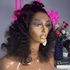 May include: A black curly human hair wig on a mannequin head. The wig is styled with loose curls and has a lace front.