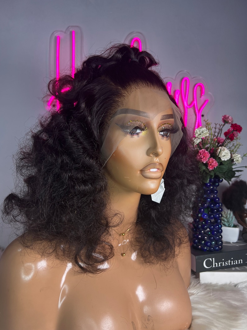 May include: A black curly human hair wig on a mannequin head. The wig is styled with loose curls and has a lace front.