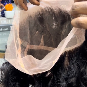May include: A close-up of a lace front wig with dark brown hair. The lace is a light pink color and is being held up to show the wig's construction.