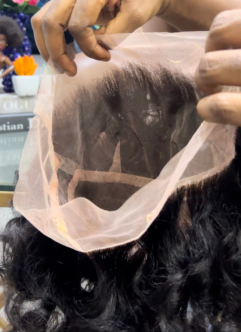 May include: A close-up of a lace front wig with dark brown hair. The lace is a light pink color and is being held up to show the wig's construction.