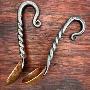 May include: Two handcrafted metal ladles with copper bowls and twisted handles. The handles are silver and curl at the top. The bowls are a warm copper color. The ladles are displayed on a dark wood surface.