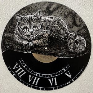 K&ouml;nnte beinhalten: Eine schwarze Schallplatte mit einer handgemalten Illustration der Grinsekatze aus Alice im Wunderland. Die Katze ist in Grau- und Wei&szlig;t&ouml;nen gehalten. Der untere Teil der Platte hat ein Zifferblatt-Design mit r&ouml;mischen Ziffern.