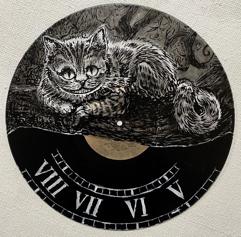 K&ouml;nnte beinhalten: Eine schwarze Schallplatte mit einer handgemalten Illustration der Grinsekatze aus Alice im Wunderland. Die Katze ist in Grau- und Wei&szlig;t&ouml;nen gehalten. Der untere Teil der Platte hat ein Zifferblatt-Design mit r&ouml;mischen Ziffern.