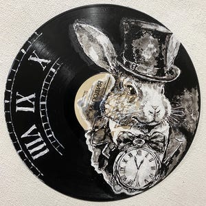 K&ouml;nnte beinhalten: Schwarze Vinyl-Schallplatte mit einem wei&szlig;en Hasen in Zylinder und Anzug, der eine Taschenuhr h&auml;lt. Der Rand der Schallplatte hat ein Zifferblatt-Design mit r&ouml;mischen Ziffern. Der Hase ist in Graustufen gemalt.