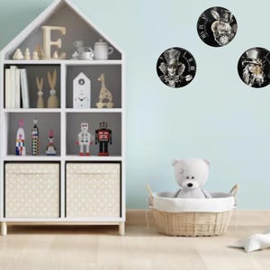 K&ouml;nnte beinhalten: Ein Kinderzimmer mit einem wei&szlig;en B&uuml;cherregal, Spielzeug und f&uuml;nf runden Uhren mit Alice im Wunderland-Charakteren. Ein hellblauer Roller und eine Baumdekoration aus Holz befinden sich links. Ein wei&szlig;er Teddyb&auml;r sitzt in einem Weidenkorb.