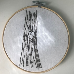Op de afbeelding: Borduurwerk in een borduurring met een boomstam met een uitgehouwen hart. Het hart heeft de initialen "J+M" erin.