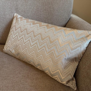 Può includere: Un cuscino rettangolare decorativo con un motivo a zig-zag dorato e grigio chiaro. Il cuscino è appoggiato su un divano marrone chiaro. Il tessuto del cuscino ha una leggera lucentezza, aggiungendo un tocco di eleganza al design.