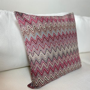 May include: Square decorative pillow with a zigzag pattern in shades of red, burgundy, blue, and beige. The pillow is resting on a white sofa, showcasing its textured design and color palette.