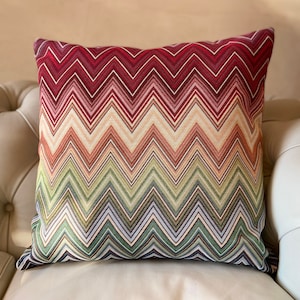 Puede incluir: Un cojín cuadrado decorativo con un vibrante patrón de chevrón. El cojín presenta un degradado de colores, incluyendo burdeos, rosa, beige, verde y azul. El cojín está colocado sobre un sofá color crema.