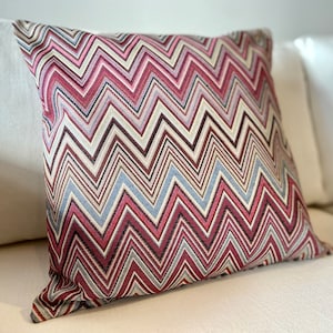 Puede incluir: Una almohada decorativa con un patrón de chevron en tonos de rosa, rojo, marrón y azul. La almohada está hecha de una tela suave y texturizada.