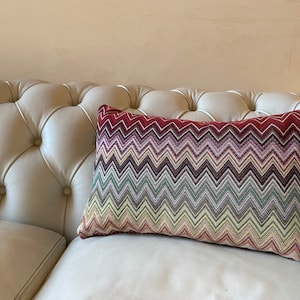 Pode incluir: Uma almofada retangular decorativa com um padrão de chevron colorido em tons de vermelho, roxo, verde e amarelo. A almofada está sobre um sofá branco de couro com tufos, criando um contraste de texturas e cores.