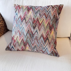 Può includere: Cuscino quadrato decorativo con un motivo a zig-zag multicolore. Il cuscino presenta una miscela di tonalità rosse, blu, gialle e verdi, creando un aspetto vibrante e strutturato. È posto su un divano bianco, suggerendo un elemento di arredo per la casa.