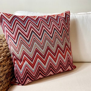 May include: A square decorative pillow with a red, white, and gray zigzag pattern. The pillow is on a white couch next to a woven basket. The pillow's design adds a pop of color and texture to the neutral setting.