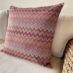 May include: A red, orange, and white chevron patterned throw pillow. The pillow is on a white couch.