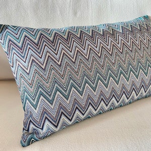 Peut inclure: Coussin rectangulaire décoratif avec un motif en zigzag bleu, blanc et marron. Le coussin est posé sur une surface blanche. Le tissu a un aspect texturé, ajoutant un intérêt visuel au design. Le coussin mesure environ 50 cm de long.