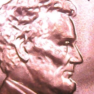 May include: A copper-colored United States one-cent coin featuring a profile portrait of Abraham Lincoln facing right. The metallic surface shows the embossed facial features, hair, and collar of the figure, with the letters US visible at the top.