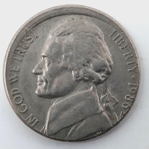 May include: A silver nickel coin featuring a profile of Thomas Jefferson, the words "LIBERTY" and "IN GOD WE TRUST", and the year 1986. The coin has a detailed design and a slightly worn appearance.