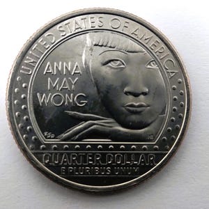 May include: A United States quarter dollar coin featuring a portrait of Anna May Wong. The coin has the words "United States of America", "Anna May Wong", and "Quarter Dollar" inscribed on it. The coin is silver in color.