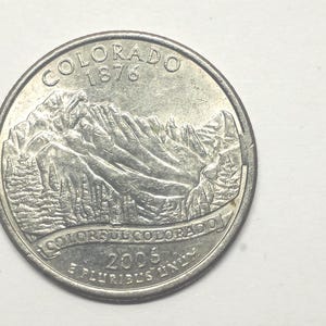 May include: A silver Colorado quarter coin. The coin features a mountain range with trees, the words "COLORADO 1876" at the top, and "COLORFUL COLORADO" and "2006 E PLURIBUS UNUM" at the bottom.