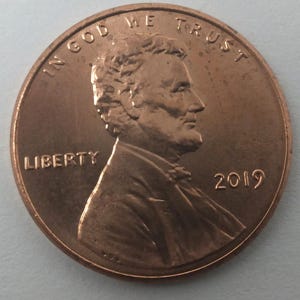 May include: A copper-colored United States penny coin. The obverse features a profile of Abraham Lincoln, the word "LIBERTY", the year "2019", and the phrase "IN GOD WE TRUST".
