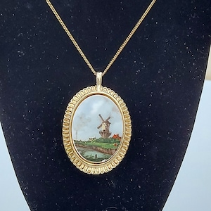 May include: A gold chain necklace with an oval pendant featuring a painted scene of a windmill in a landscape. The pendant has a gold frame with a beaded edge.