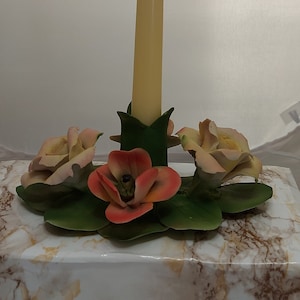 May include: A white marble candle holder with a single yellow candle and three porcelain flowers. Two of the flowers are peach colored and one is orange.