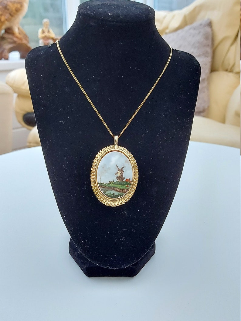 May include: A gold chain necklace with an oval pendant featuring a painted scene of a windmill in a landscape. The pendant has a gold frame with a beaded edge.
