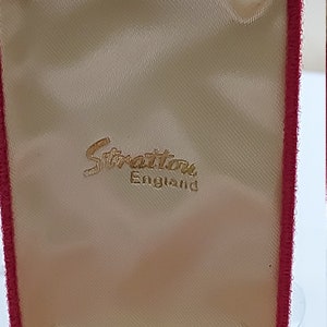 May include: A red velvet jewelry box with a cream satin lining. The box has a gold embossed label that reads "Stratton England".