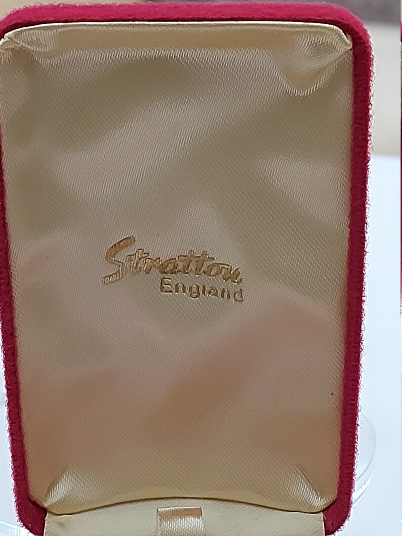 May include: A red velvet jewelry box with a cream satin lining. The box has a gold embossed label that reads "Stratton England".