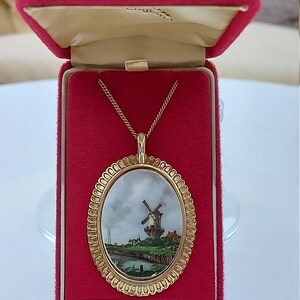 May include: A gold-plated oval pendant necklace with a painted scene of a windmill in a landscape. The pendant is in a red velvet box with the words "Guaranteed Gold Plated" printed on the bottom.