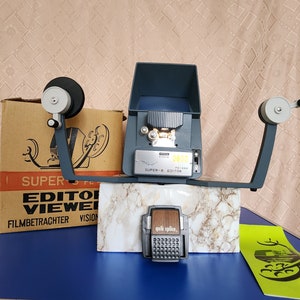 Może przedstawiać: Winylowy edytor filmu Super 8 z wbudowanym wizjerem, parą srebrnych ramion i czarno-brązowym splicerom do taśmy filmowej "quik splice". Edytor stoi na marmurowej powierzchni z brązowym pudełkiem i zielonym papierem z logo jelenia w tle.