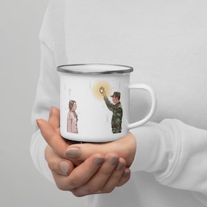 Puede incluir: Taza de esmalte blanco con un gráfico de una persona con uniforme militar sosteniendo una vela encendida y dándosela a una persona con cabello castaño corto. El fondo es un patrón de rayas azul claro y rosa.