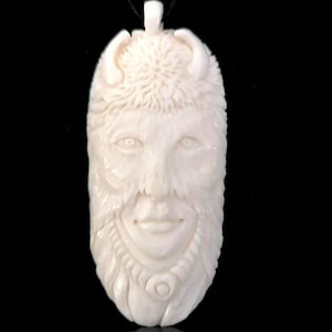 Può includere: Un ciondolo bianco intagliato che raffigura un viso con corna e dettagli intricati. Il ciondolo è fatto di osso e pende da un cordoncino nero.