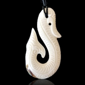 May include: A white bone carved fish hook pendant with intricate details, hanging from a black cord. The hook is a traditional Maori design, known as a Hei Matau.