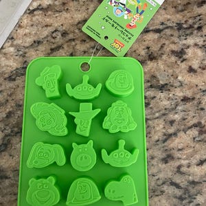Könnte beinhalten: Eine grüne Silikon-Schokoladenform mit verschiedenen Toy Story-Charakterdesigns. Die Form enthält Bilder von Woody, Buzz Lightyear und anderen Charakteren. Ein grünes Etikett mit japanischem Text ist angebracht.