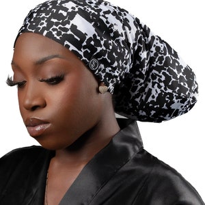 May include: A black and white patterned satin bonnet. The bonnet features a repeating abstract design and a small button detail. The person in the image is wearing a black satin robe and a gold necklace.