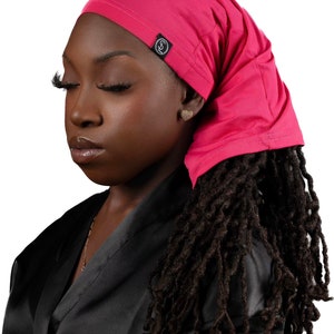 May include: A woman with dreadlocks is wearing a pink satin head wrap with a black logo. She is wearing a black satin robe.