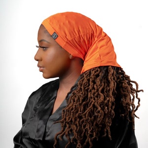 Könnte beinhalten: Eine Frau mit langen braunen Dreadlocks trägt eine leuchtend orangefarbene Satin-Schlafhaube. Die Haube hat ein kleines schwarzes Logo an der Seite.