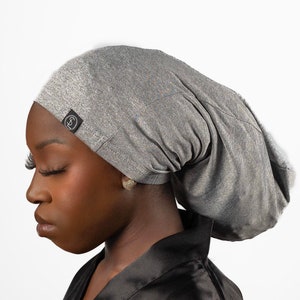 May include: A gray satin sleep cap with a small black logo on the side. The cap is worn by a person with dark skin and is styled to cover the hair completely.