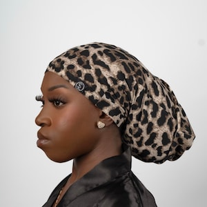 Puede incluir: Una persona lleva una gorra de dormir de satén con estampado de leopardo marrón y negro. La gorra está recogida en la parte superior y tiene un pequeño logotipo negro en un lateral.