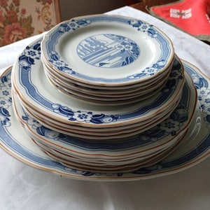 Peut inclure: Une pile d'assiettes en céramique blanche avec des motifs floraux et paysagers bleus. Les assiettes ont un bord festonné et une bordure bleue avec un fin accent marron. Les assiettes sont empilées sur une nappe blanche.