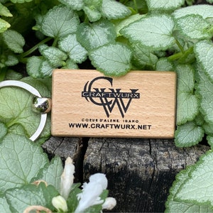 May include: A wooden keychain tag with the Craft Wurx logo and website address. The tag is rectangular and light-colored, with black text. A silver keyring is attached. The background features green and silver foliage.