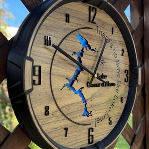 May include: A round wooden wall clock featuring a blue resin inlay of Lake Coeur d'Alene. The clock face has black numerals, a compass rose icon, and the text "There's no time like Lake time!" with black metal hands and a black frame.